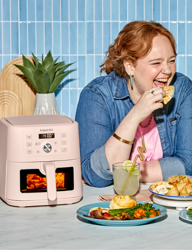 A woman with red hair, wearing a denim shirt and pink top, laughs while eating a snack at a table set with plates of food and a drink. Next to her is the Instant Pot® 4QT VORTEX® Plus Mini Air Fryer in Rosewater, with its front window showing food cooking inside.