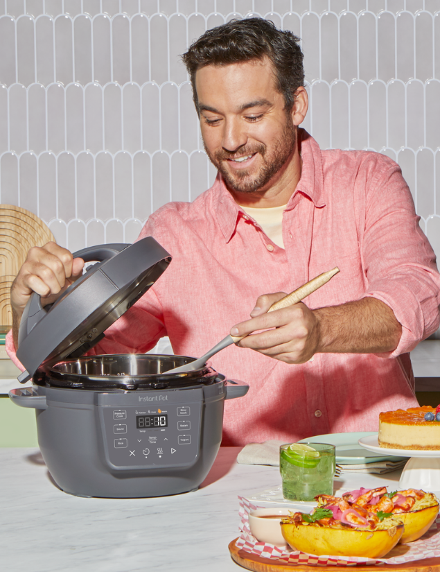 A man in a coral shirt smiles while stirring food inside the Instant Pot® RIO™ Mini 4QT Multi-Cooker in Truffle, with the lid open. A slice of cheesecake, stuffed squash, and a green drink sit on the table in front of him.