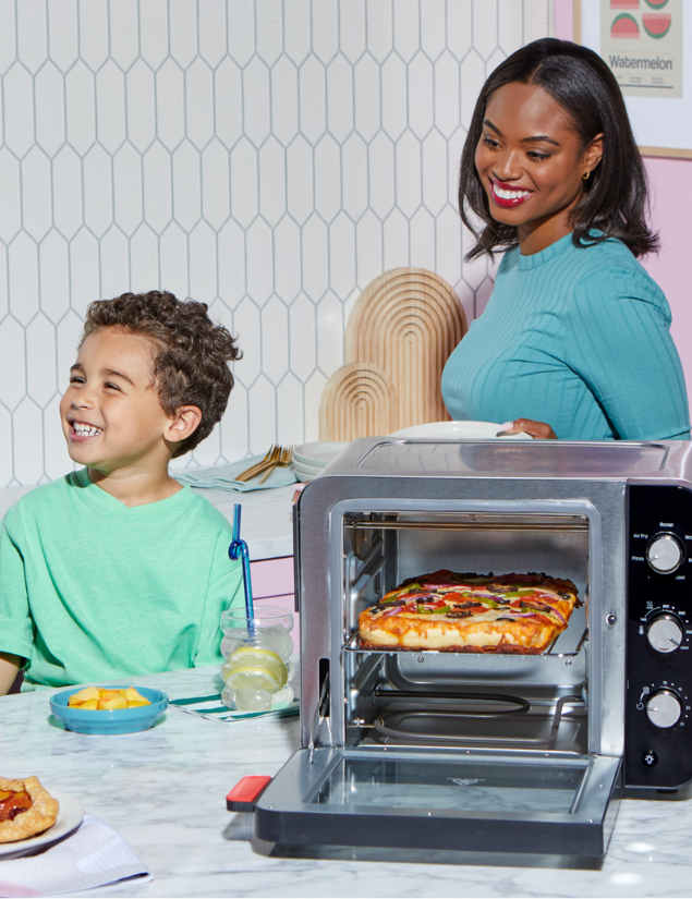 A woman in a teal top stands beside a young boy in a matching teal shirt, both smiling at the table. In front of them, a pizza bakes inside the Instant Pot® 4-Slice InstantHeat™ Mini Air Fryer Toaster Oven in Stainless Steel. A small plate of fruit and a drink are also on the table.