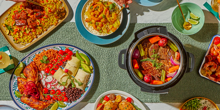 Overhead view of a colorful spread of home-cooked meals on a bright table. At the center, an Instant Pot filled with tender pot roast, carrots, celery, red onions, and potatoes. Surrounding dishes include a platter of grilled chicken with rice, black beans, tamales, avocado slices, and cherry tomatoes; a bowl of golden shrimp curry over rice; a plate of glazed salmon with couscous and vegetables; and a tray of shrimp boil with corn and potatoes. Fresh limes, herbs, and gold utensils add vibrant accents.