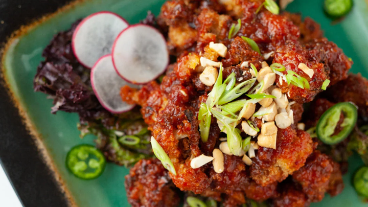 A close-up of crispy cauliflower bites coated in a glossy red spicy sauce, topped with chopped peanuts and sliced green onions, served on a green plate with sliced jalapeños and radishes on the side.