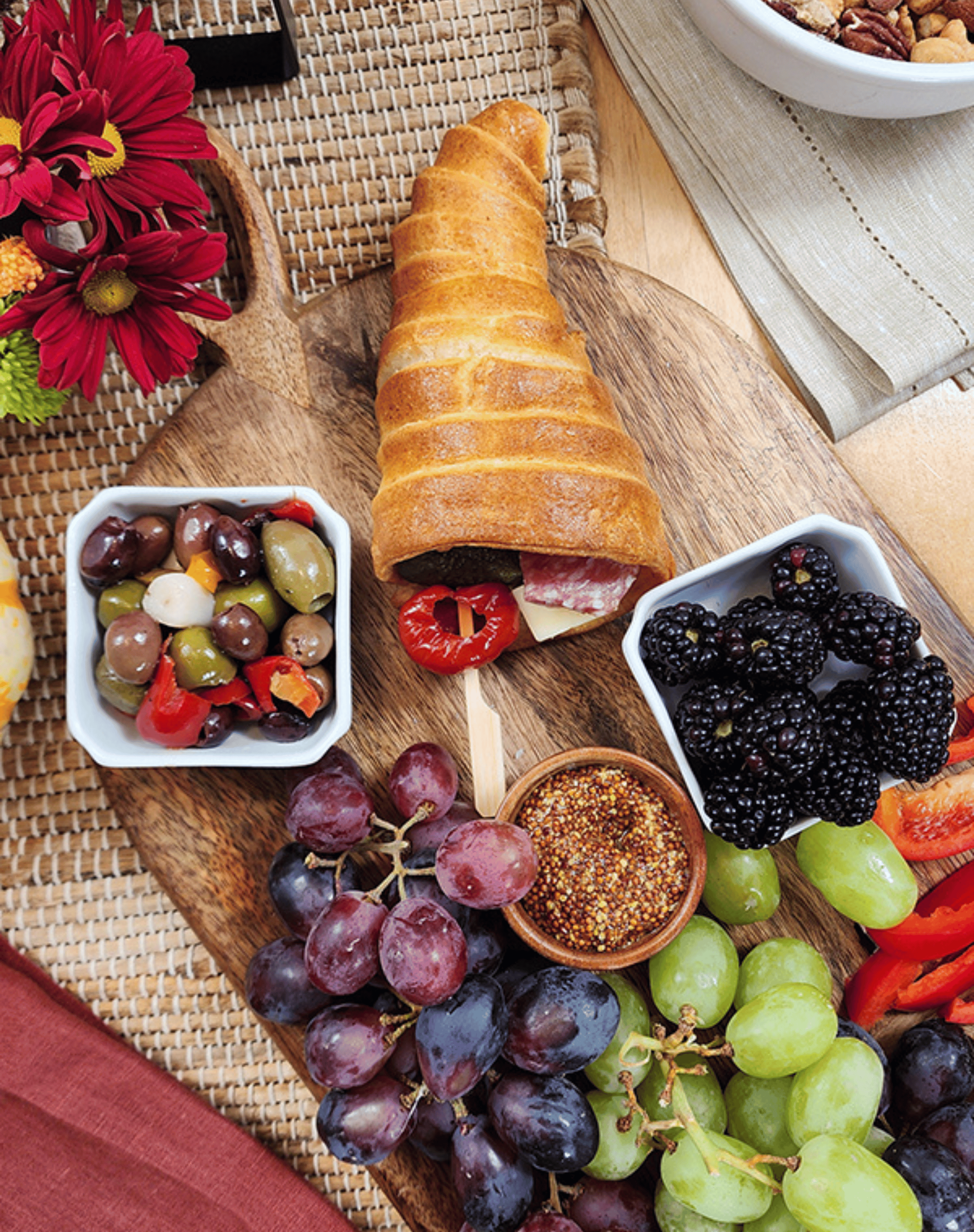 A festive charcuterie cornucopia filled with slices of salami, cheese, and stuffed peppers spilling from a golden crescent roll horn onto a wooden board. Surrounding it are small bowls of marinated olives, grainy mustard, and fresh blackberries, along with clusters of red and green grapes. The warm wood tones and autumn flowers add a cozy, harvest-inspired feel.