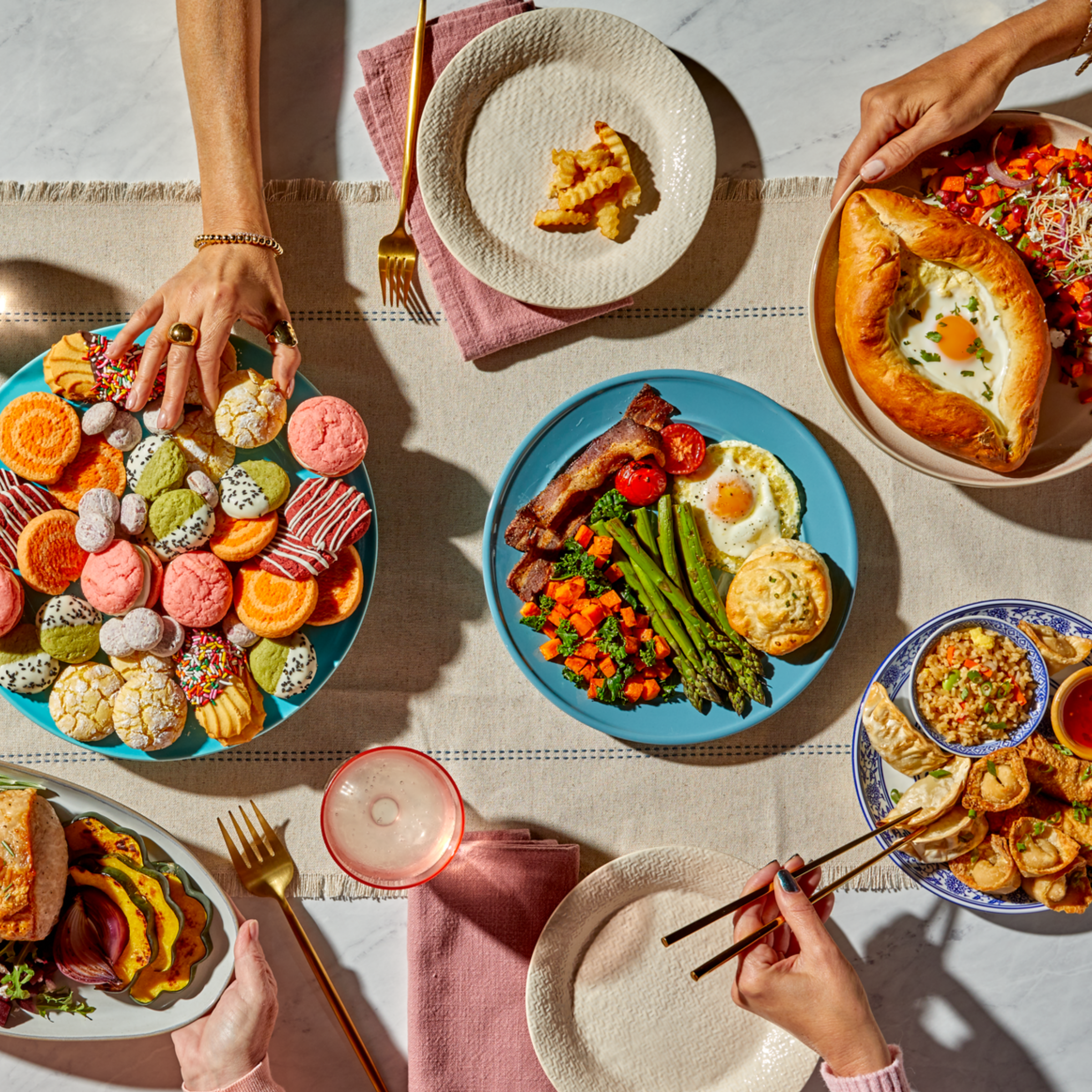 Overhead view of a colorful shared meal with various dishes, including cookies, breakfast with eggs and bacon, khachapuri, dumplings, fried rice, and a sandwich with roasted vegetables. Multiple hands reach in, creating a lively, communal dining scene.