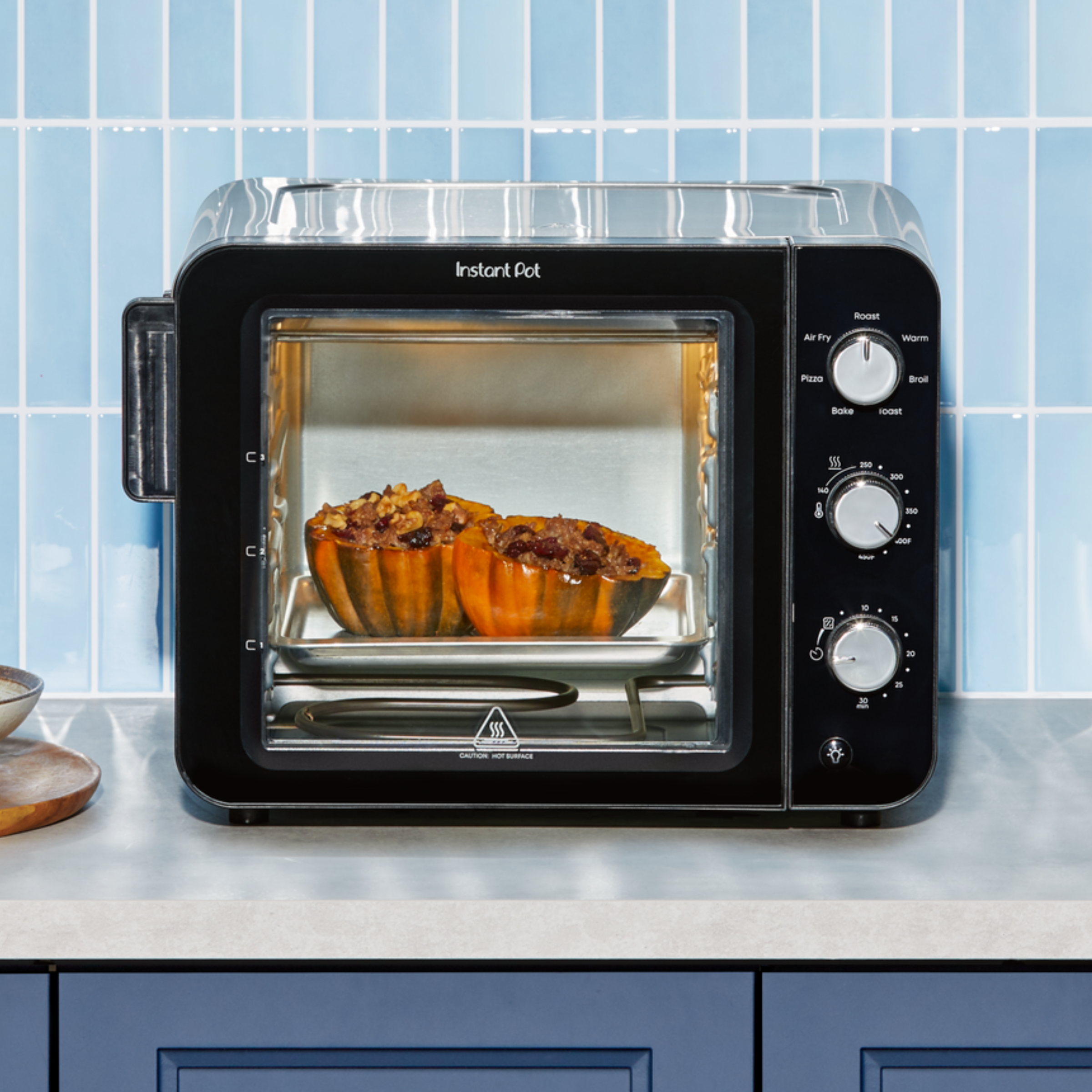 A front-facing view of the Instant Pot® 4-Slice InstantHeat™ Mini Air Fryer and Toaster Oven in black, placed on a light gray kitchen counter with a blue tiled backsplash. The oven door is closed, and the interior light reveals two halved acorn squashes stuffed with a savory filling roasting inside. The appliance features three chrome dials on the right for selecting cooking functions, temperature (up to 450°F), and time. 