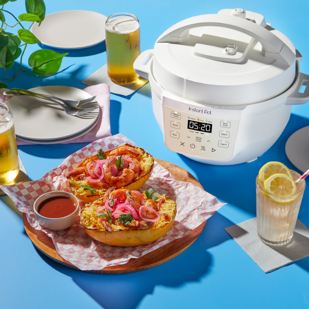 The Instant Pot® 4QT RIO™ Mini Multi-Cooker sits on a bright blue table, displaying a cooking time of 5:20 on its digital screen. In front of the appliance, a wooden tray holds two spaghetti squash boats topped with sauce, pickled onions, and herbs. The table is set with drinks, plates, and utensils, creating a vibrant, inviting mealtime scene.