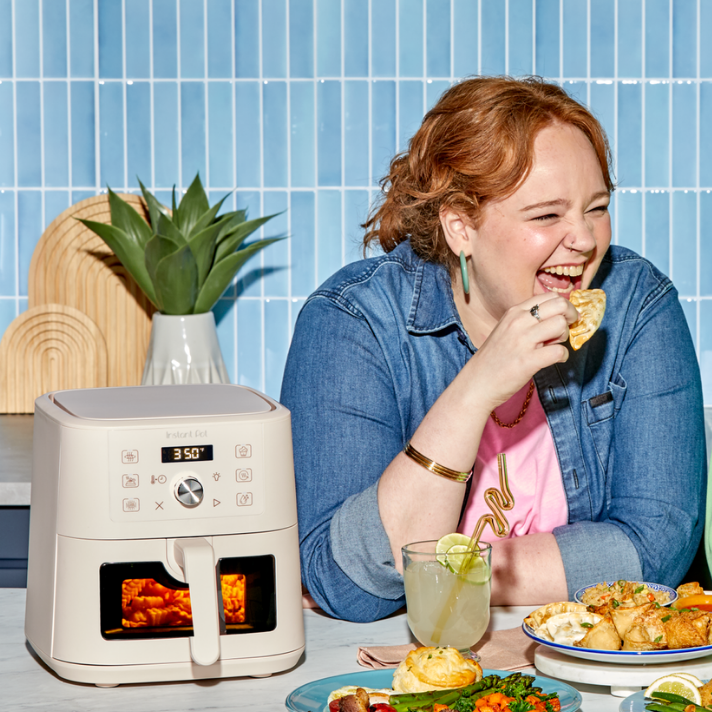 A joyful woman with wavy red hair, wearing a denim shirt over a pink tee with a squiggle design, laughs as she takes a bite of a crispy, golden turnover. In front of her on the kitchen counter is the Instant Pot® 4QT VORTEX® Plus Mini Air Fryer in a soft Rosewater color, glowing warmly through its clear front window as food cooks inside. The appliance has a digital display reading “350°F” and icons for various cooking functions. Surrounding her are vibrant plates of food.