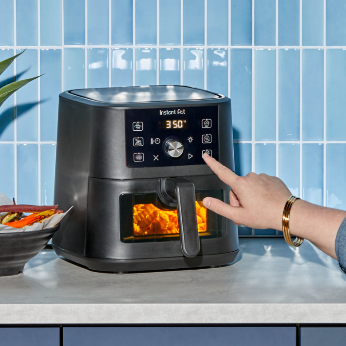 A close-up of a hand pressing a button on the digital control panel of the Instant Pot® 4QT VORTEX® Plus Mini Air Fryer in sleek Black. The air fryer is set to 350°F, with multiple cooking icons lit up on the display, including air fry and bake. Through the clear window on the drawer-style basket, golden food is visibly crisping under the bright internal light. The appliance sits on a light gray kitchen counter with a bowl of colorful veggie chips nearby, against a glossy light blue tile backsplash.