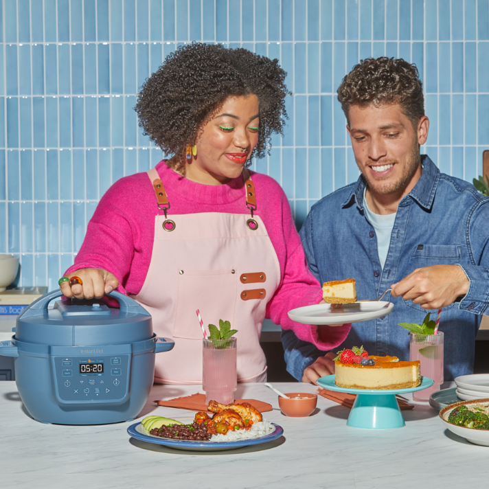 A woman in a bright pink sweater and pale pink apron smiles as she holds the lid handle of a blue Instant Pot® 4QT RIO™ Mini Chef Series Multi-Cooker in Daydream. Beside her, a man in a denim shirt serves a slice of cheesecake onto a plate, smiling. The countertop is set with colorful, inviting dishes, including a plate of shrimp, rice, black beans, and avocado, a cheesecake topped with berries and orange glaze, and two tall pink drinks garnished with mint and striped straws. 