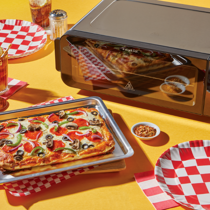 The Instant Pot® InstantHeat™ Pizza and Toaster Oven sits on a bright yellow table, reflecting a fresh rectangular pizza that rests on a baking tray nearby. The scene is set for a casual meal with red-and-white checkered plates, drinks, and small bowls of red pepper flakes and dipping sauce.