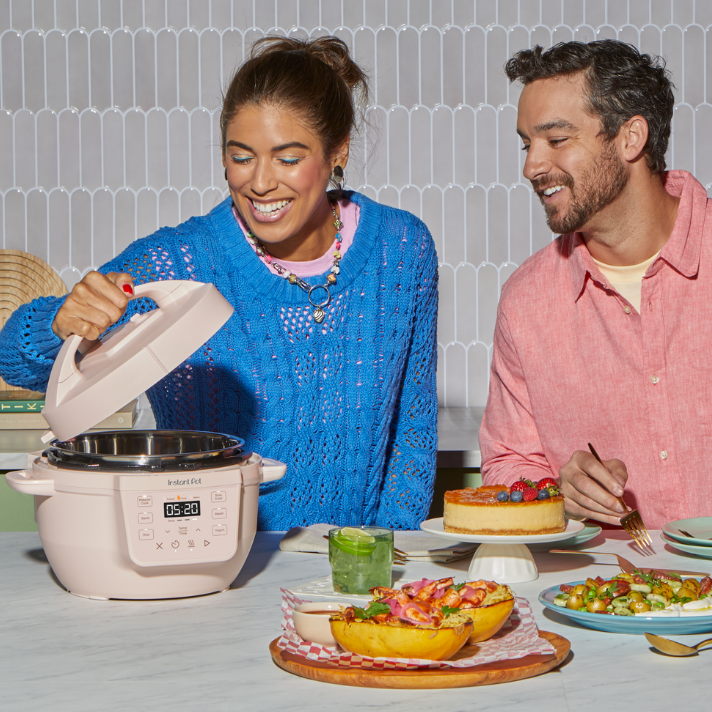 A woman in a bright blue sweater smiles as she opens the lid of a Rosewater-colored Instant Pot® 4QT RIO™ Mini Multi-Cooker on a kitchen counter. A man in a pink shirt sits beside her, smiling while holding a fork and a plate with cheesecake topped with berries. The counter is set with vibrant, colorful dishes including stuffed bread rolls, a green drink, and a vegetable salad. The scene is cheerful and casual, showcasing the product in a home cooking setting.