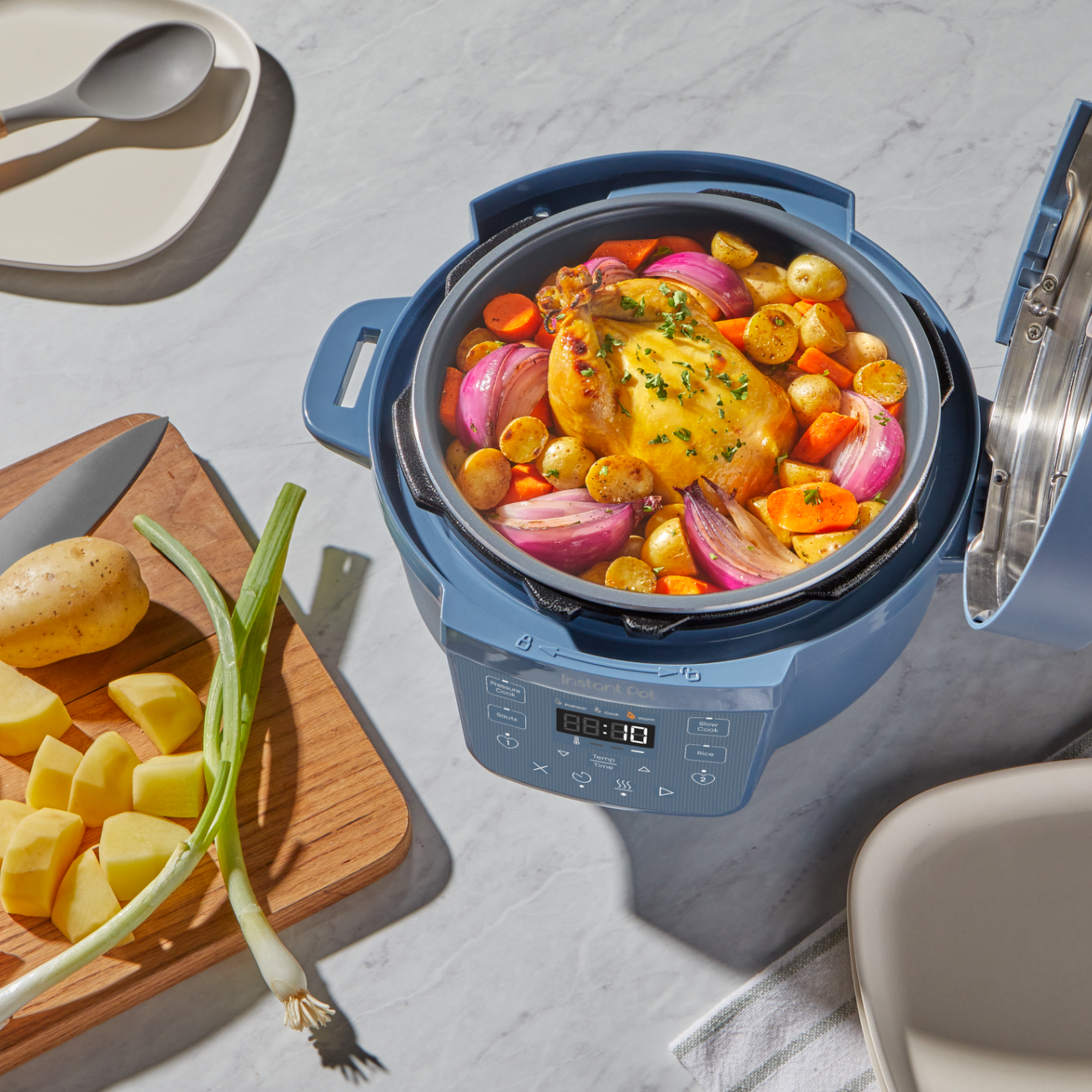 The Instant Pot® 4QT RIO™ Mini Chef Series Multi-Cooker in Daydream sits on a marble countertop with its lid open, revealing a whole roasted chicken surrounded by carrots, baby potatoes, and red onions. Nearby, a cutting board holds chopped vegetables and a green onion, capturing a cozy, home-cooked meal in progress.