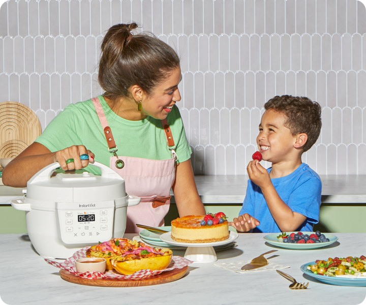 A woman and young boy smile at each other in a bright kitchen while enjoying fresh food. In front of them is the Instant Pot® 4QT RIO™ Mini Multi-Cooker in Sea Salt, surrounded by colorful dishes including cheesecake, stuffed squash, berries, and a veggie pasta salad.