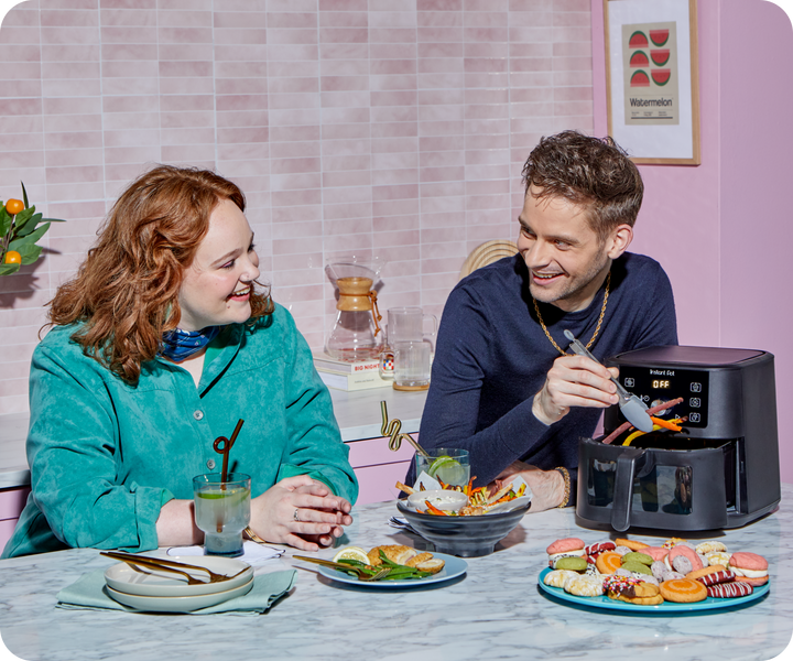 Two people laugh and enjoy snacks in a colorful kitchen while using the Instant Pot® 4QT VORTEX® Plus Mini Air Fryer in Black, which is open with fresh fries inside. The marble counter is covered with vibrant dishes, including spring rolls, dipping sauces, and a platter of assorted cookies and pastries.