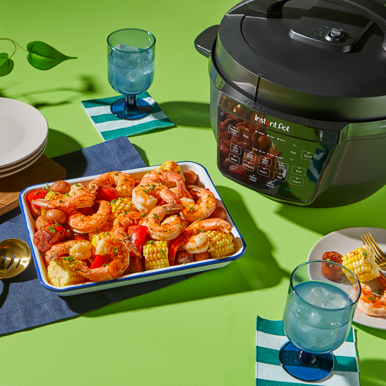A vibrant seafood boil featuring shrimp, corn on the cob, red potatoes, and bell peppers is served in a white tray with blue trim, placed next to an Instant Pot with a sleek black exterior and wide control panel. The scene is set on a bright green table with blue-striped napkins, blue goblets filled with ice water, and white plates, highlighting the Instant Pot's spacious wide base—perfect for cooking and serving generous, crowd-pleasing meals.